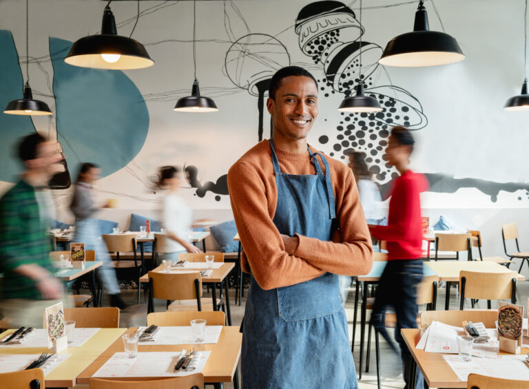 portrait of smiling male owner or waiter in cafe with unrecognizable blurred employes moving.